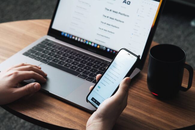 hombre tiene celular en la mano derecha y notebeook en su mesa con taza de cafe