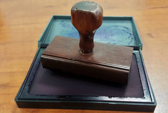 Un sello de madera oscura descansa sobre una almohadilla de tinta abierta, dentro de una caja de plástico verde oscuro sobre una superficie de madera.