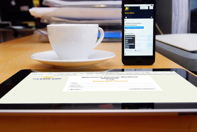 Foto-1 Una taza de café descansa sobre una mesa de madera entre una tablet y un teléfono inteligente que muestran una página web. Al fondo hay documentos apilados y una impresora."