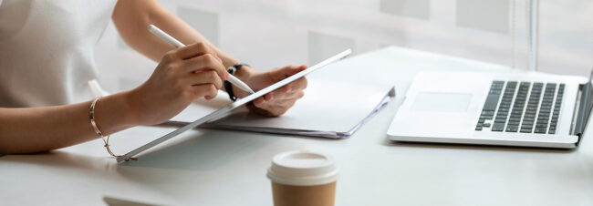 Una mujer usa un lápiz digital para interactuar con una tableta, situada sobre una mesa junto a un portátil y una taza de café.