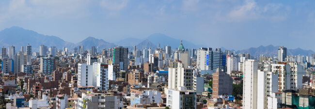 Una extensa ciudad se extiende sobre un paisaje montañoso, con edificios de diversos tamaños y alturas. Los edificios altos destacan sobre una densa colección de estructuras más bajas.