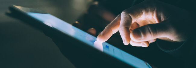 Un dedo índice interactúa con una pantalla táctil iluminada de una tablet, en un entorno oscuro.