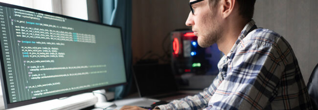 Un hombre programa en un monitor grande con código en la pantalla, sentado en un escritorio de casa con una torre de computadora cerca.