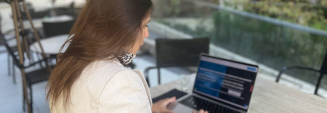 Una mujer con el pelo largo y castaño usa un notebook en una terraza al aire libre.