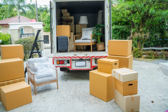 Boxes waiting to be moved into a new home,New home,Moving house day and real estate concept. Un camión de mudanzas, cargado con muebles y cajas, está estacionado en una entrada. Varias cajas adicionales esperan ser cargadas. El contexto es un patio residencial con vegetación.