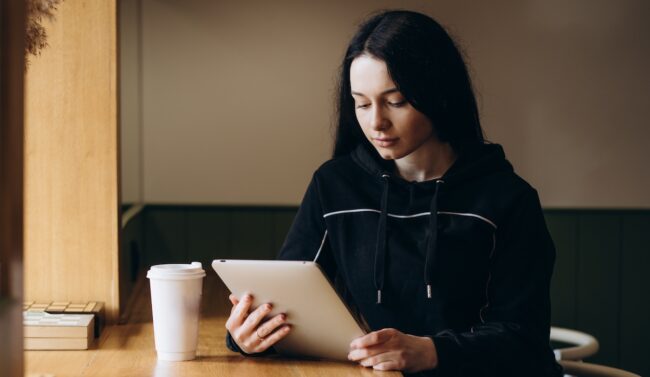 Una joven, sentada en un café, mira atentamente una tablet digital que sostiene en sus manos. Un vaso de bebida se encuentra sobre la mesa junto a ella.