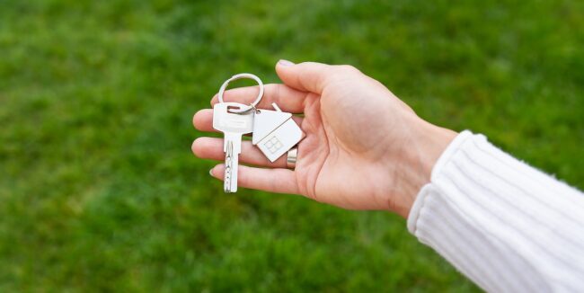 Una mano sostiene un llavero con llaves y un pequeño dije de casa. El llavero se muestra sobre un fondo de césped verde.