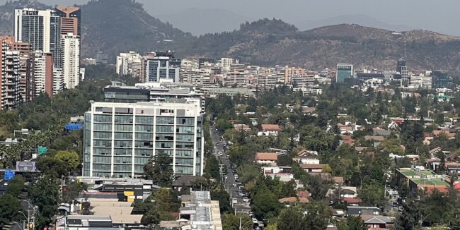 Un edificio de oficinas de varios pisos se encuentra en un barrio residencial densamente arbolado, en las laderas de una colina, con vista a una gran ciudad y montañas al fondo.