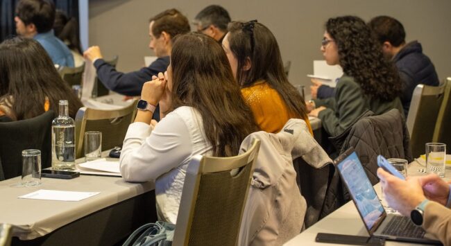 calidad de vida Un grupo de personas, la mayoría mujeres, se sientan en mesas escuchando atentamente en una sala de conferencias. Tienen papeles y ordenadores notebooks delante.