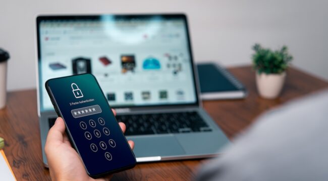 Un teléfono inteligente muestra una pantalla de inicio de sesión de seguridad; una persona introduce un código PIN. Se encuentra junto a un notebook con una página web abierta, en una mesa de escritorio.