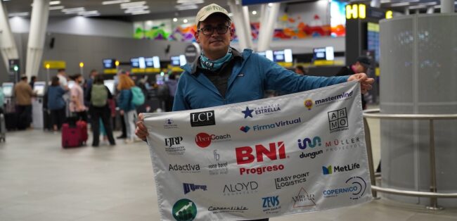 Un hombre (Christian Rodiek) sostiene un estandarte con múltiples logotipos, incluyendo "BNI Progreso", en un aeropuerto concurrido.