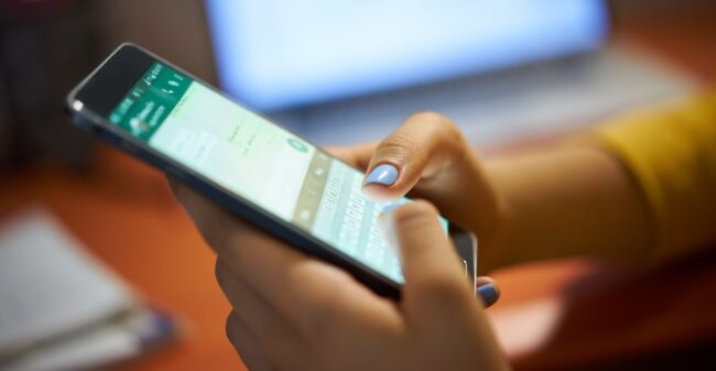 Un teléfono inteligente muestra una pantalla con una aplicación de mensajería; dos manos escriben un mensaje; el teléfono se sostiene en un escritorio, con un ordenador notebook borroso en el fondo.