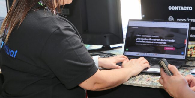 Una mujer usa un notebook mostrando una página web sobre firmas electrónicas. Sus manos teclean en el notebook; otra mano sujeta un teléfono móvil. El contexto es un puesto de trabajo o feria.