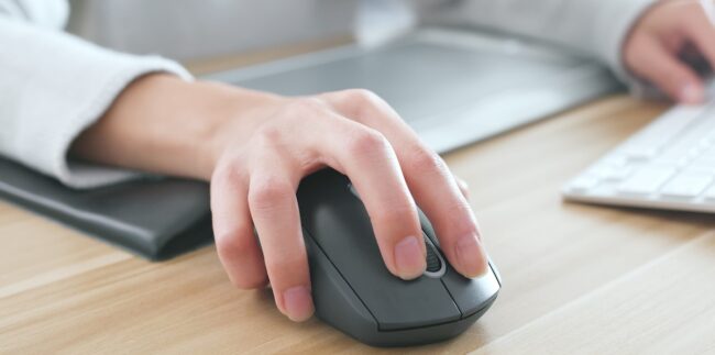 Una mano manipula un ratón de computadora gris oscuro sobre una superficie de madera. Un teclado y una almohadilla para ratón son visibles en segundo plano.