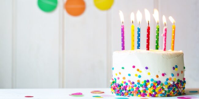 Un pastel blanco decorado con chispas de colores y ocho velas encendidas, descansa sobre una mesa blanca delante de un fondo de madera blanca con guirnaldas borrosas de círculos de colores.