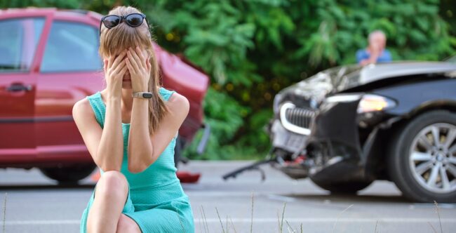 Una mujer, sentada en el suelo, cubre su rostro con las manos tras un accidente de tráfico. Un auto rojo y otro negro dañado están cerca.