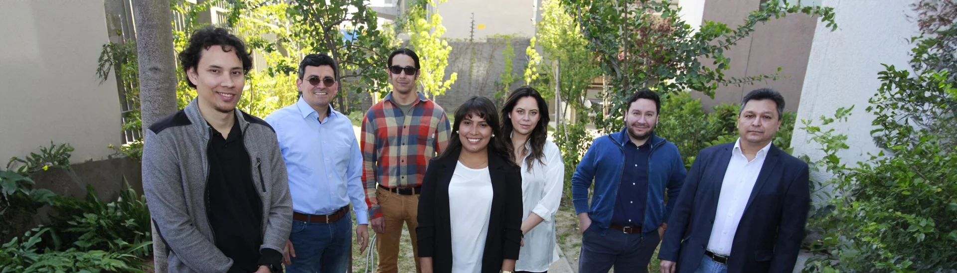 Ocho personas, posiblemente un equipo, posan juntos al aire libre entre exuberante vegetación. Están de pie, mirando hacia la cámara. El contexto es un entorno exterior con arbustos y edificios.