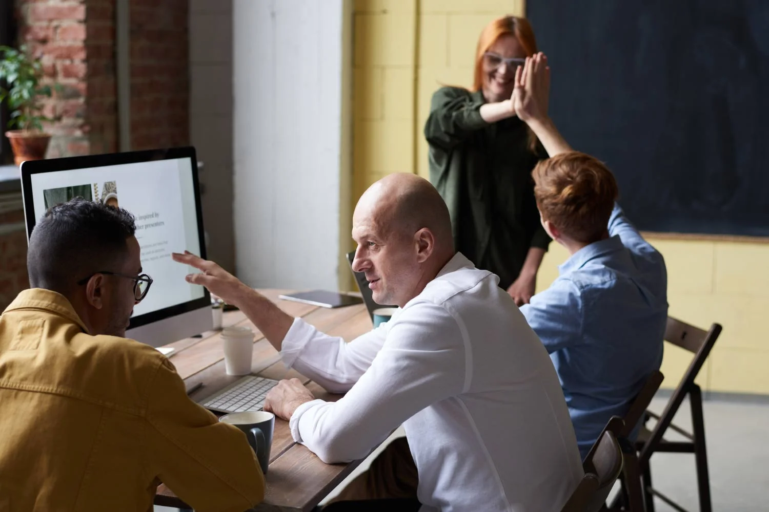 Un equipo de cuatro personas trabaja en una computadora en una oficina. Un hombre señala la pantalla mientras un compañero mira. Otra persona celebra en segundo plano.