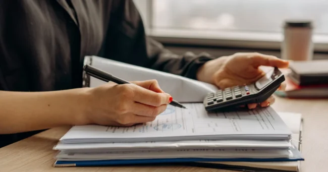 Manos con un bolígrafo y una calculadora sobre documentos, junto a una ventana
