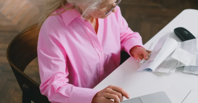 Mujer mayor revisa documentos en papel junto a un computador portátil