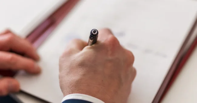 Mano de un hombre de traje firmando un documento sobre una carpeta